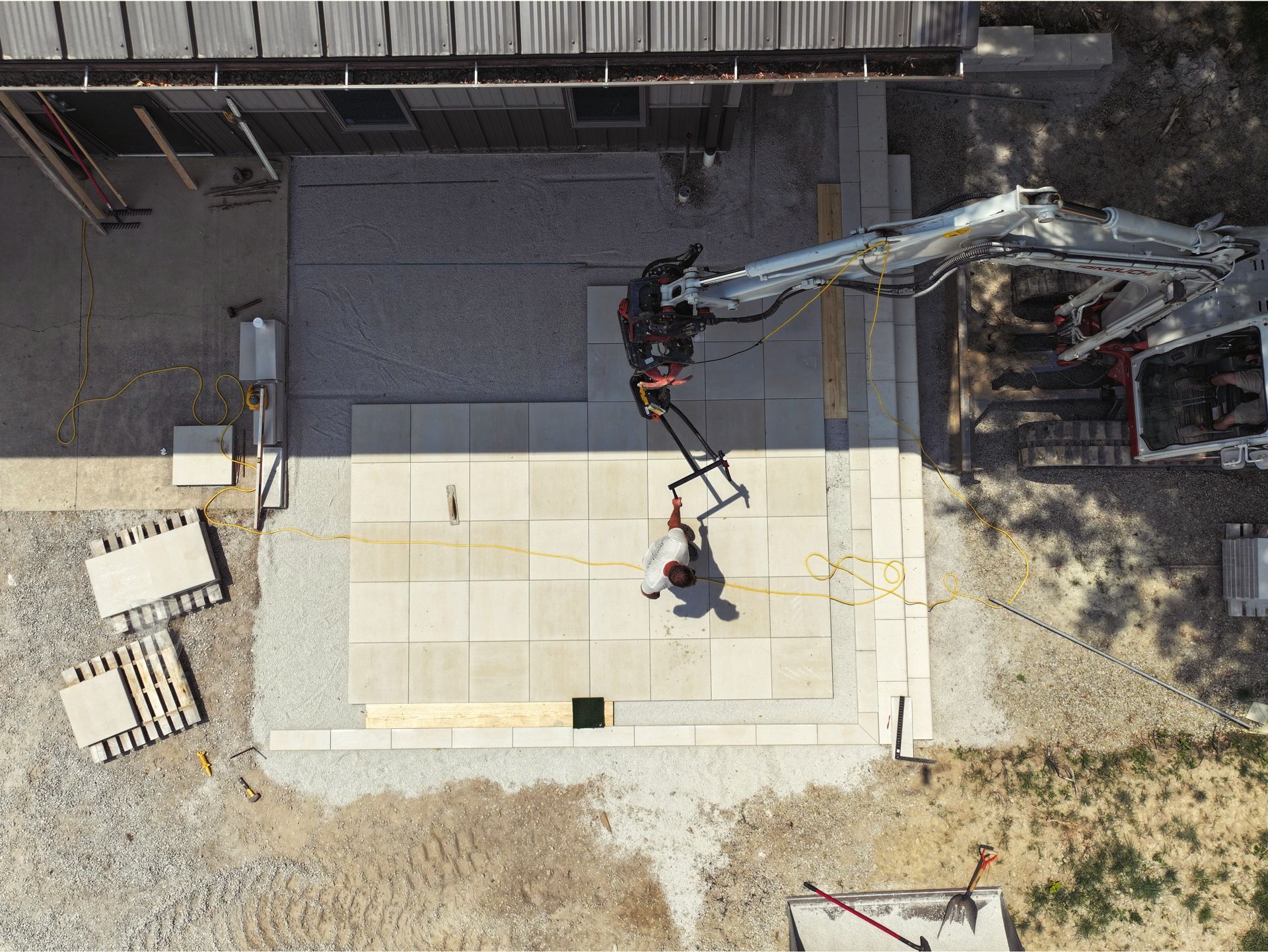 Overhead view of a construction site: a worker stands near a small excavator laying large square tiles on a patio.