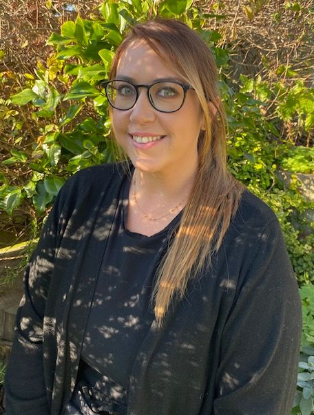 Woman with glasses smiling outdoors, wearing black cardigan, near greenery.