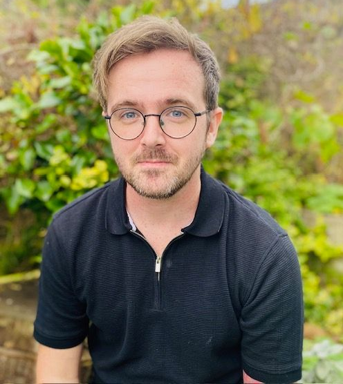 Man with glasses and a black polo shirt, smiling slightly outdoors.