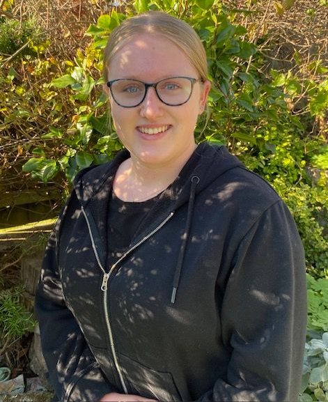 Woman with glasses smiling, wearing black hoodie, standing outdoors in front of green foliage.