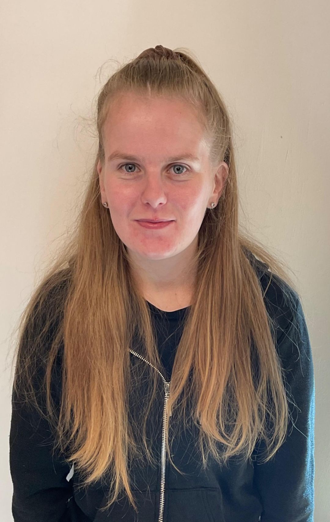 Portrait of a person with long blonde hair wearing a dark top, standing against a beige wall.