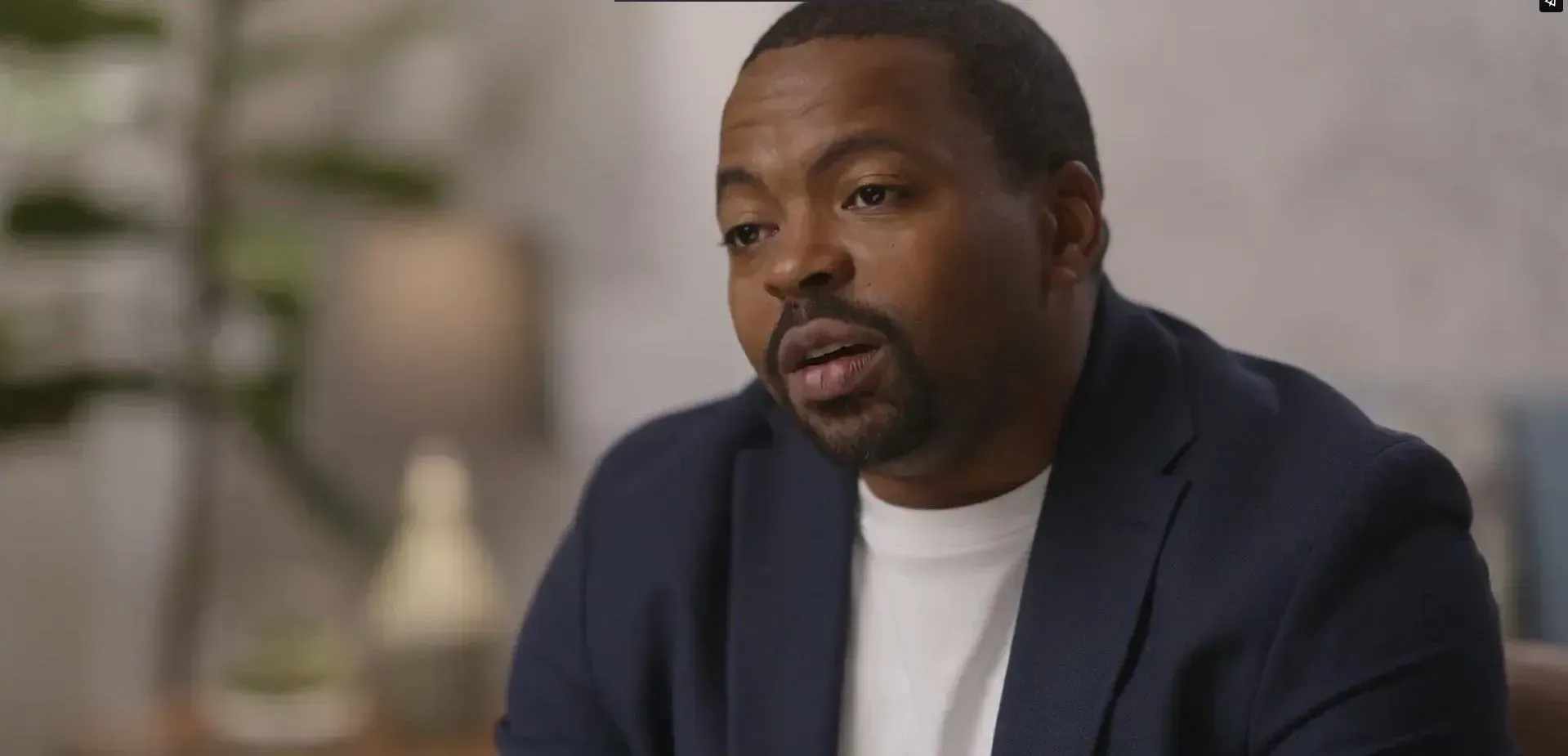 A Black man in a blue blazer and white shirt speaks in an indoor setting.