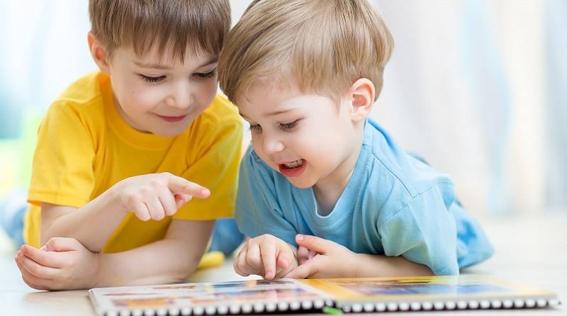 Two young children, one in yellow and one in blue, look at a book together, pointing at a page.