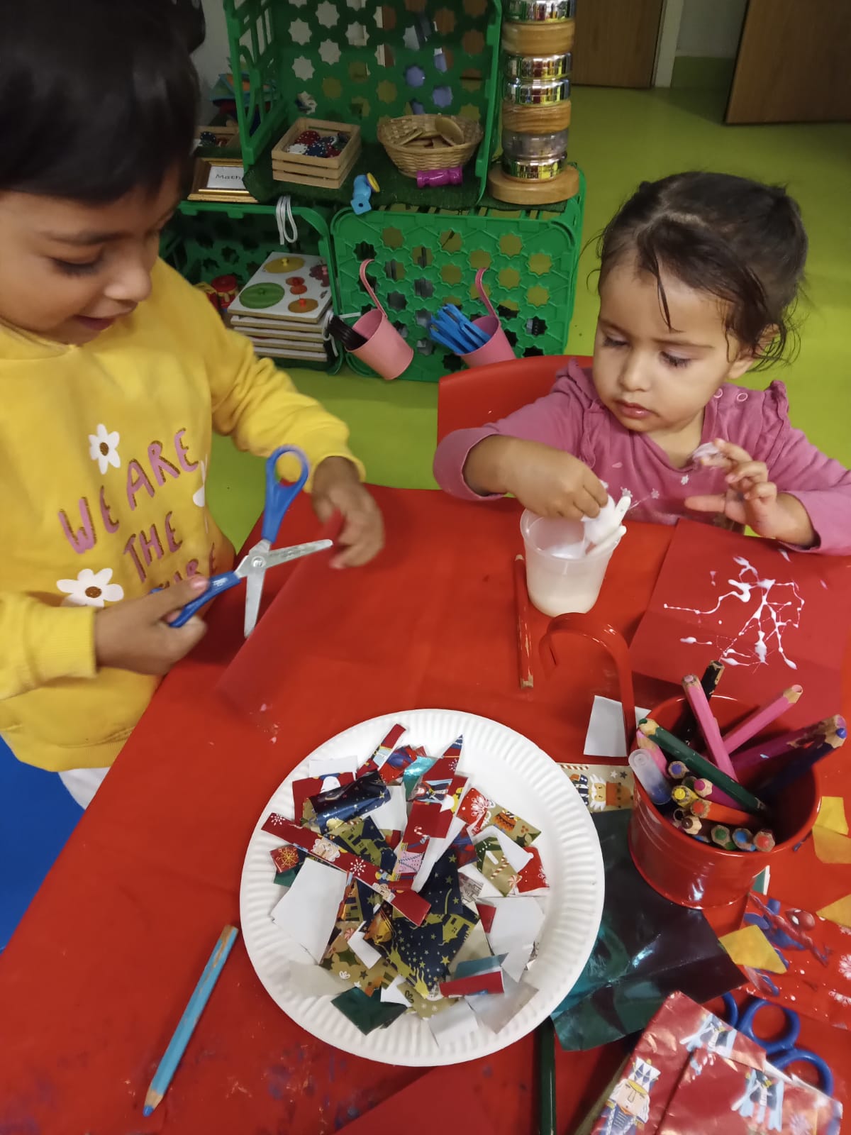 Two children at a table, cutting paper with scissors and glueing. Red table, art supplies, and colorful backdrop.