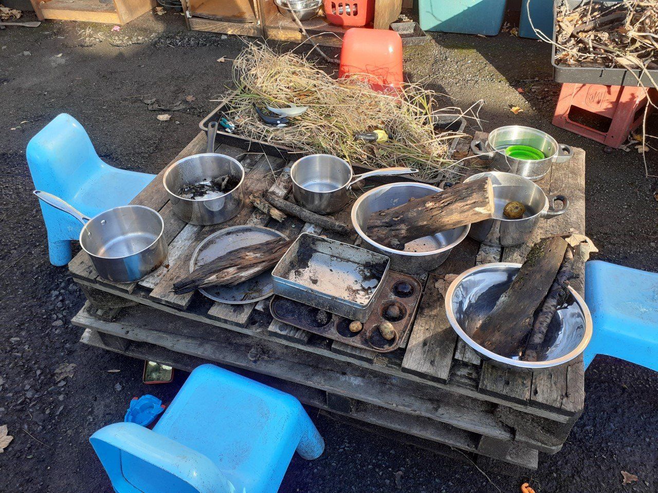 Wooden pallet with metal bowls, sticks, and green liquid, used as a play table with blue plastic chairs.
