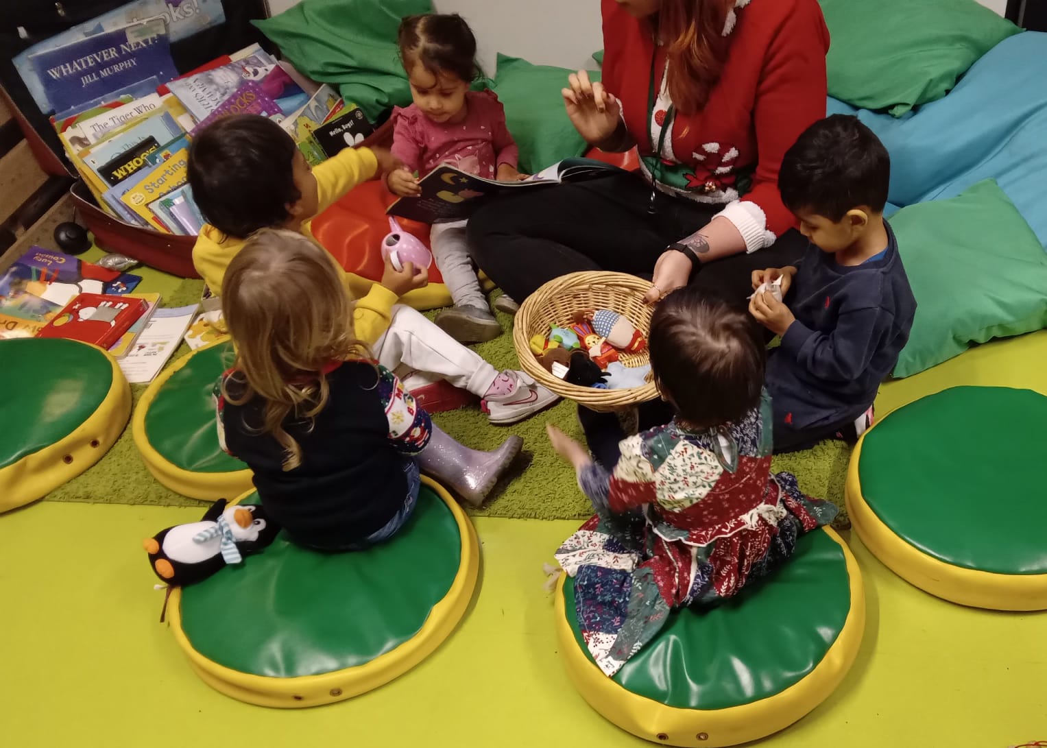 Children seated in a circle with a teacher, holding a basket of toys in a colorful playroom.