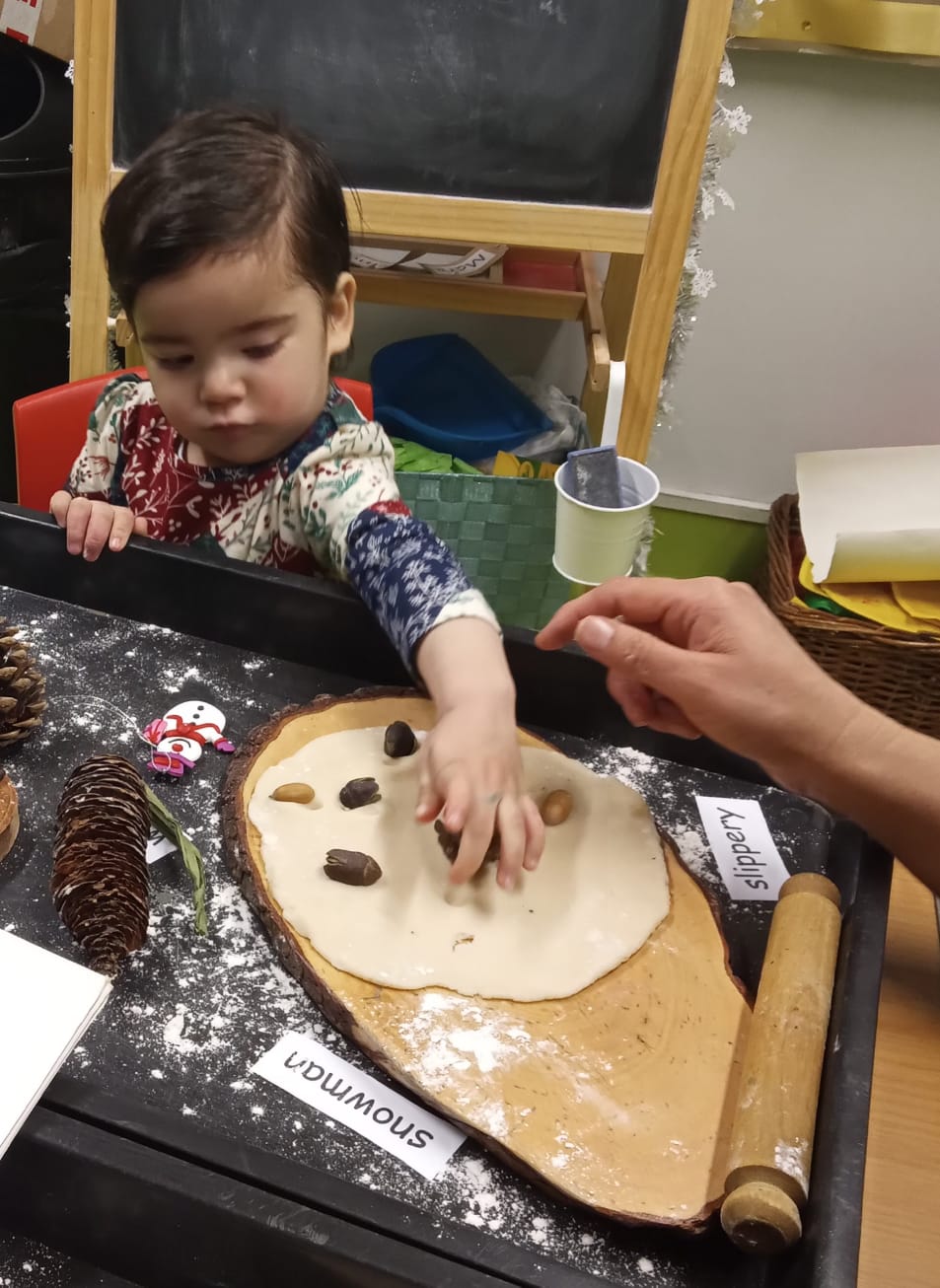 Child interacting with natural objects and a dough on a tray, with an adult's hand nearby.