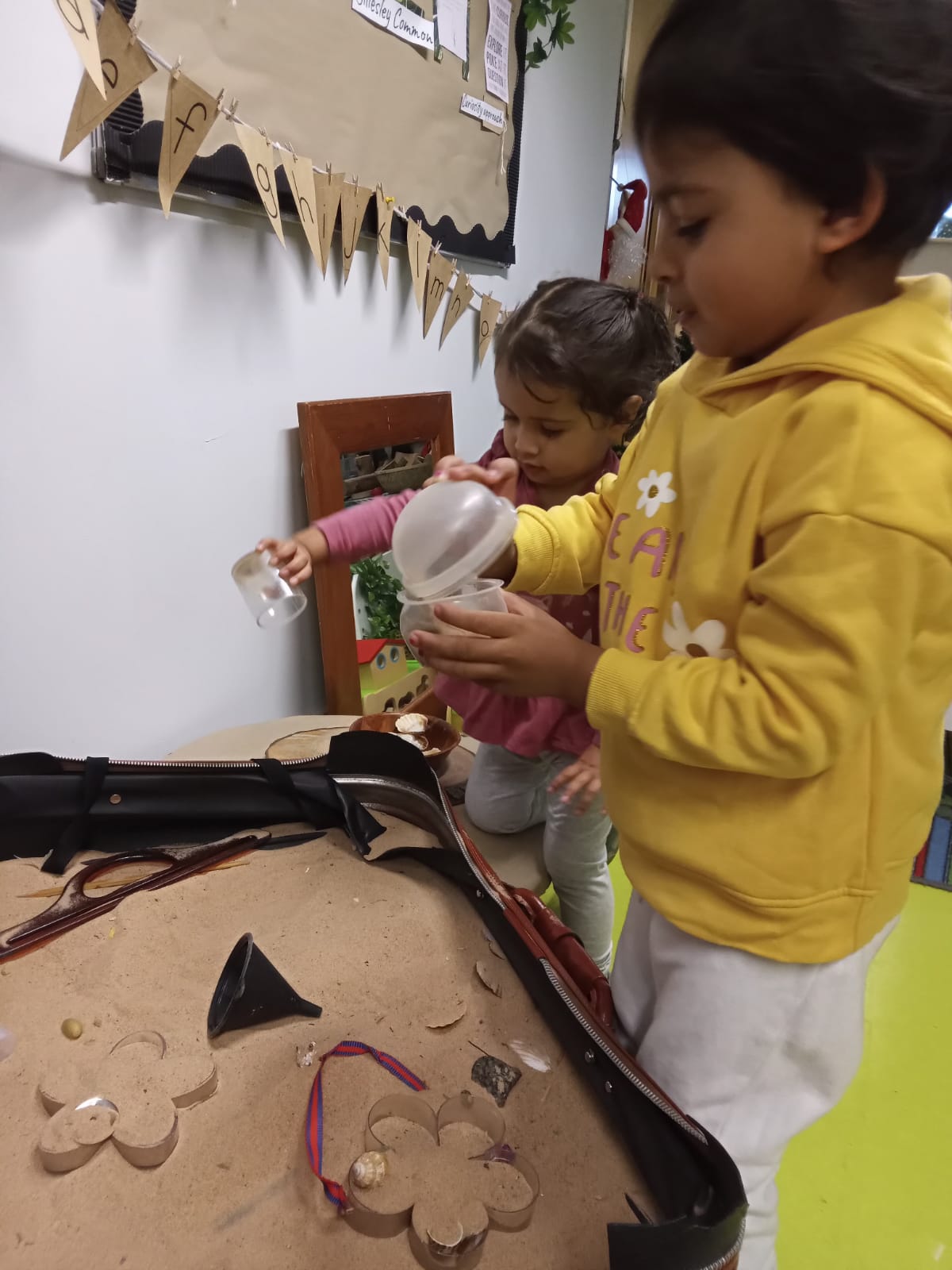 Two children playing in a sand pit, one pouring from a cup.