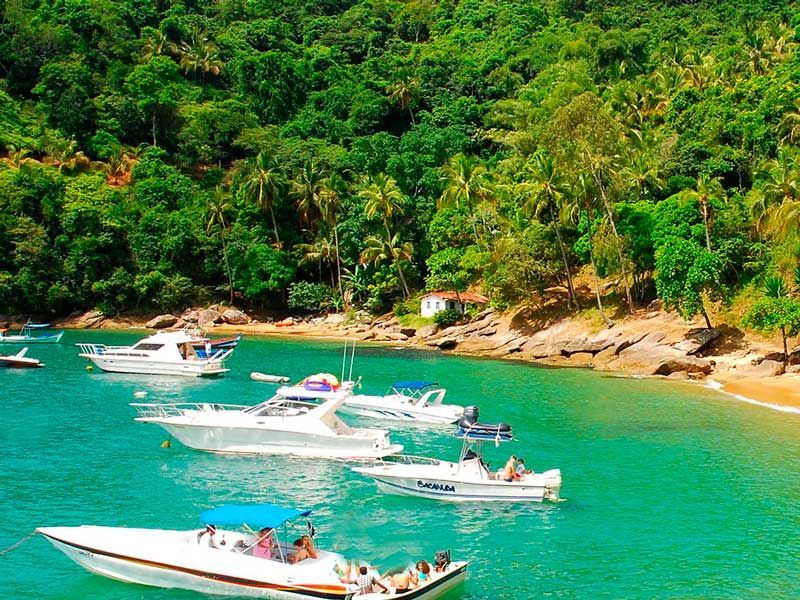Diversas lanchas flutuam em águas azul-turquesa brilhantes perto de uma praia de areia rodeada por uma densa floresta tropical verde.