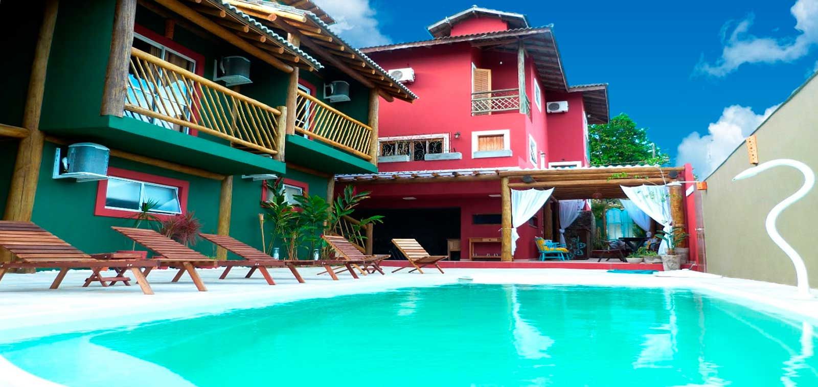 Uma piscina azul brilhante fica em frente a um prédio verde de dois andares e a uma casa vermelha de vários andares com espreguiçadeiras.