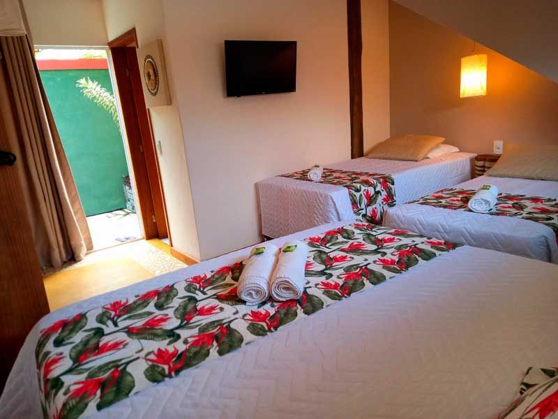 Interior de um quarto de hotel com duas camas de solteiro com lençóis brancos, caminhos de mesa florais, toalhas brancas enroladas e uma TV fixada na parede.