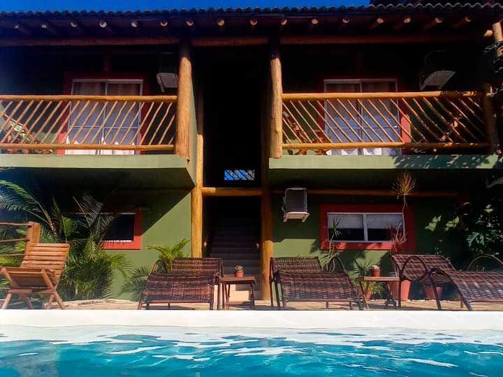 Um chalé verde de dois andares com varandas e espreguiçadeiras, situado ao lado de uma piscina de água cristalina azul.