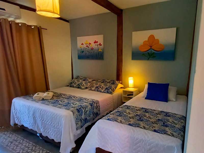 Um quarto com uma cama de casal e uma cama de solteiro, com peseiras estampadas, dois quadros na parede e iluminação aconchegante ao lado da cama.