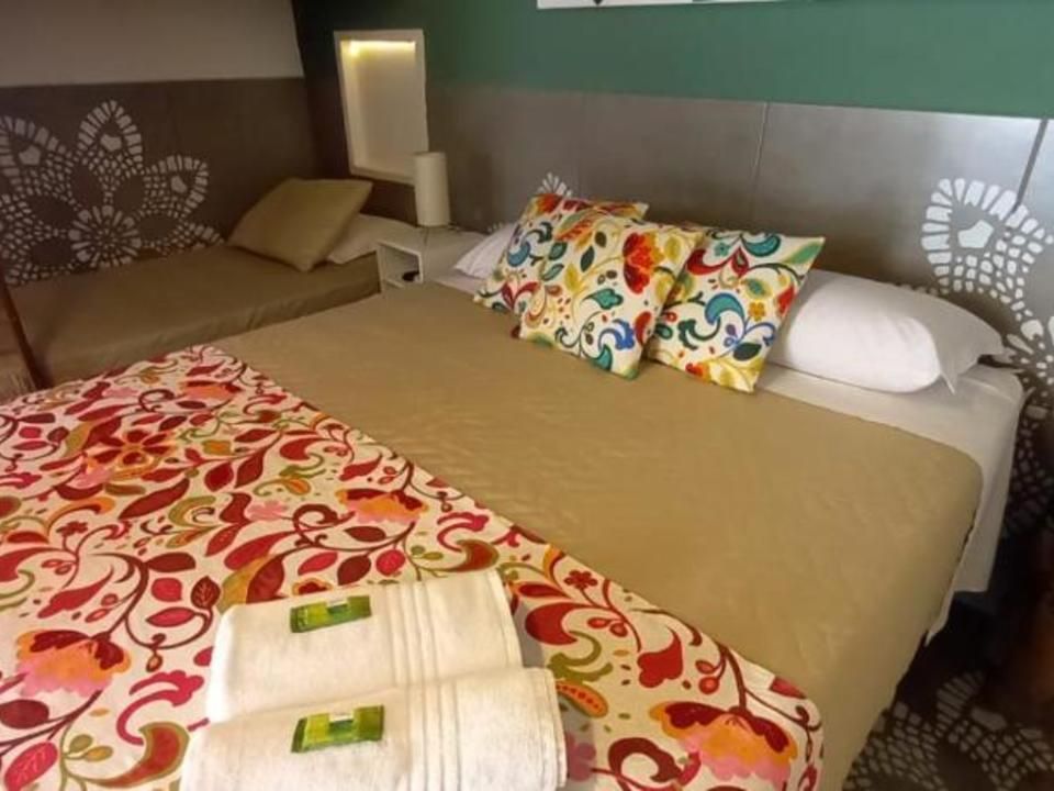 Um quarto de hotel com uma cama de casal impecavelmente arrumada, com um caminho de mesa floral, duas almofadas estampadas e toalhas brancas dobradas.