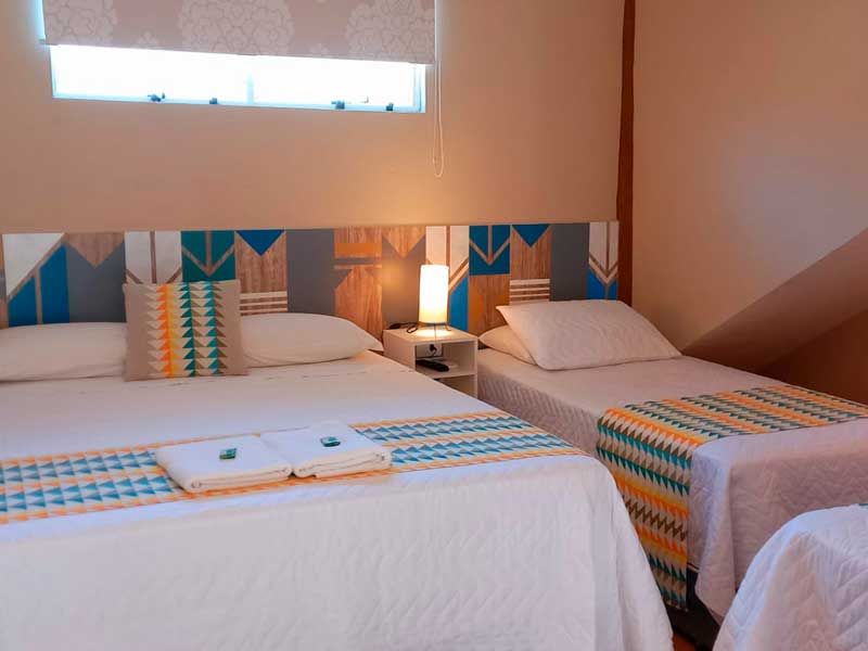 Um quarto com duas camas de lençóis brancos, caminhos de mesa estampados e uma cabeceira geométrica colorida com um pequeno abajur.
