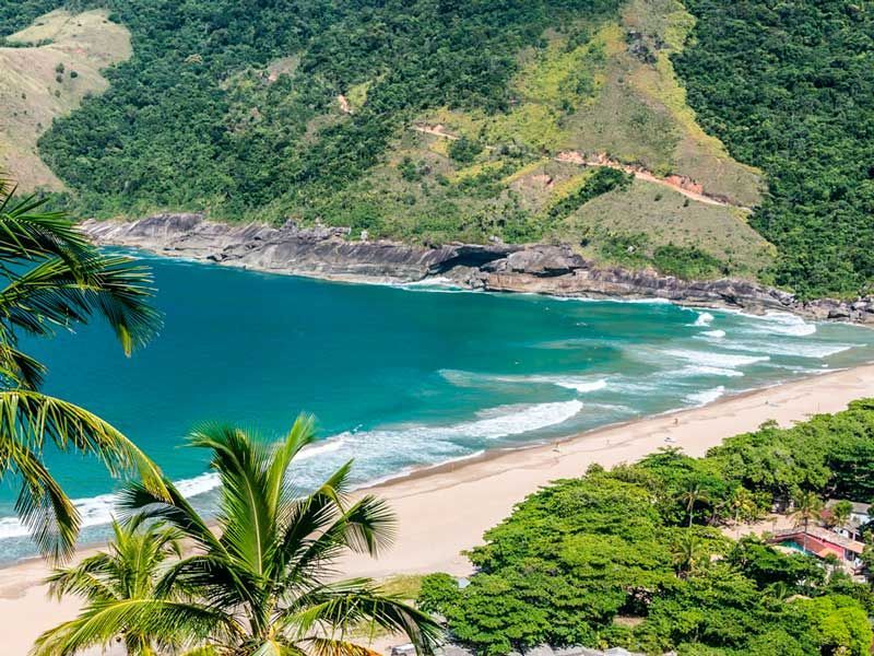 Vista aérea de uma praia tropical de areia com água turquesa, falésias rochosas escuras e floresta verdejante.