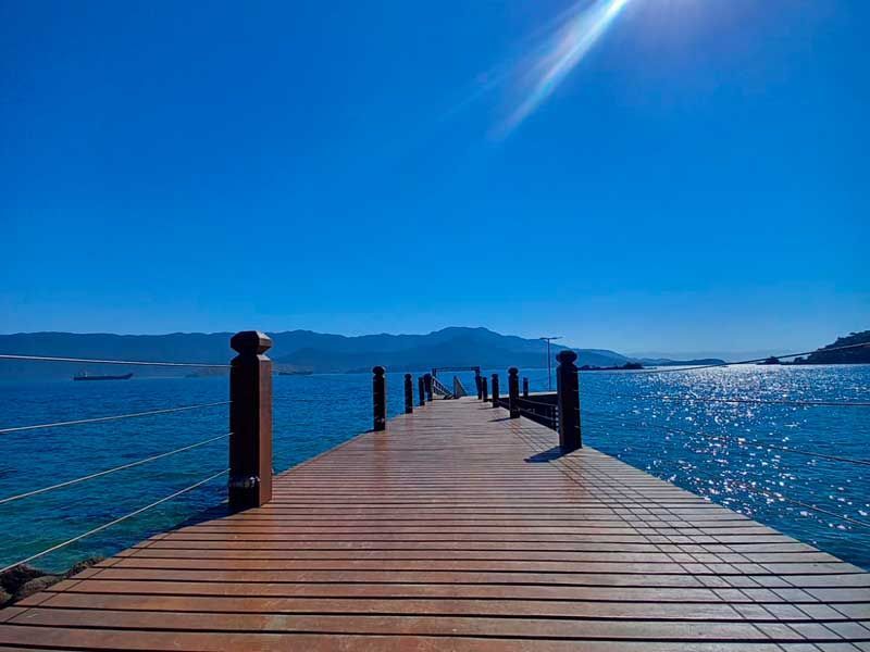 Um cais de madeira estende-se por um mar calmo e azul profundo, sob um céu claro e brilhante, com montanhas distantes no horizonte.