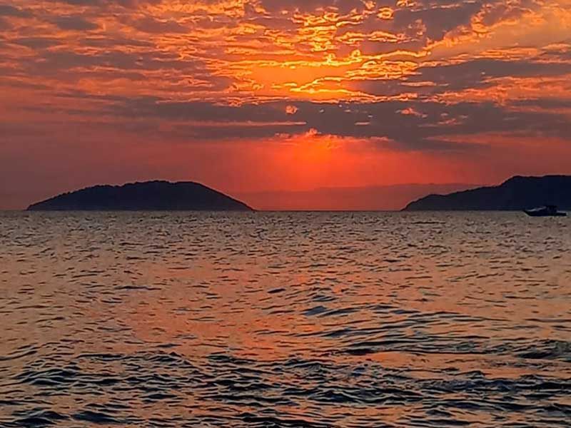 Um pôr do sol alaranjado vibrante ilumina um mar calmo, com duas ilhas silhuetadas ao longe e um pequeno barco à direita.