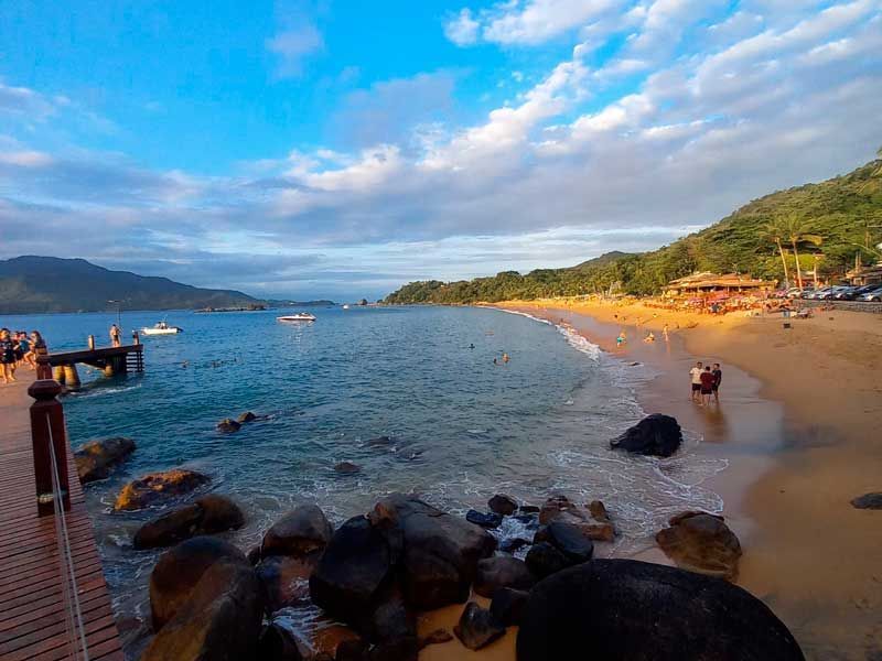Uma paisagem costeira com um cais de madeira que se estende pelas águas azuis, uma praia de areia, rochas escuras e colinas verdejantes.