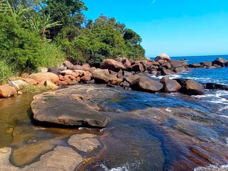 Uma costa rochosa com grandes pedras lisas e marrons encontrando as águas cristalinas do oceano azul sob um céu ensolarado e brilhante.