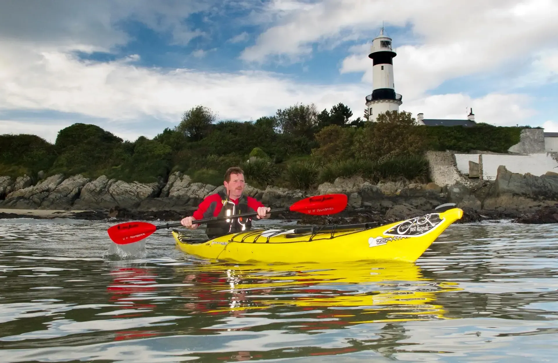 A man is paddling a yellow kayak in the water with a lighthouse in the background