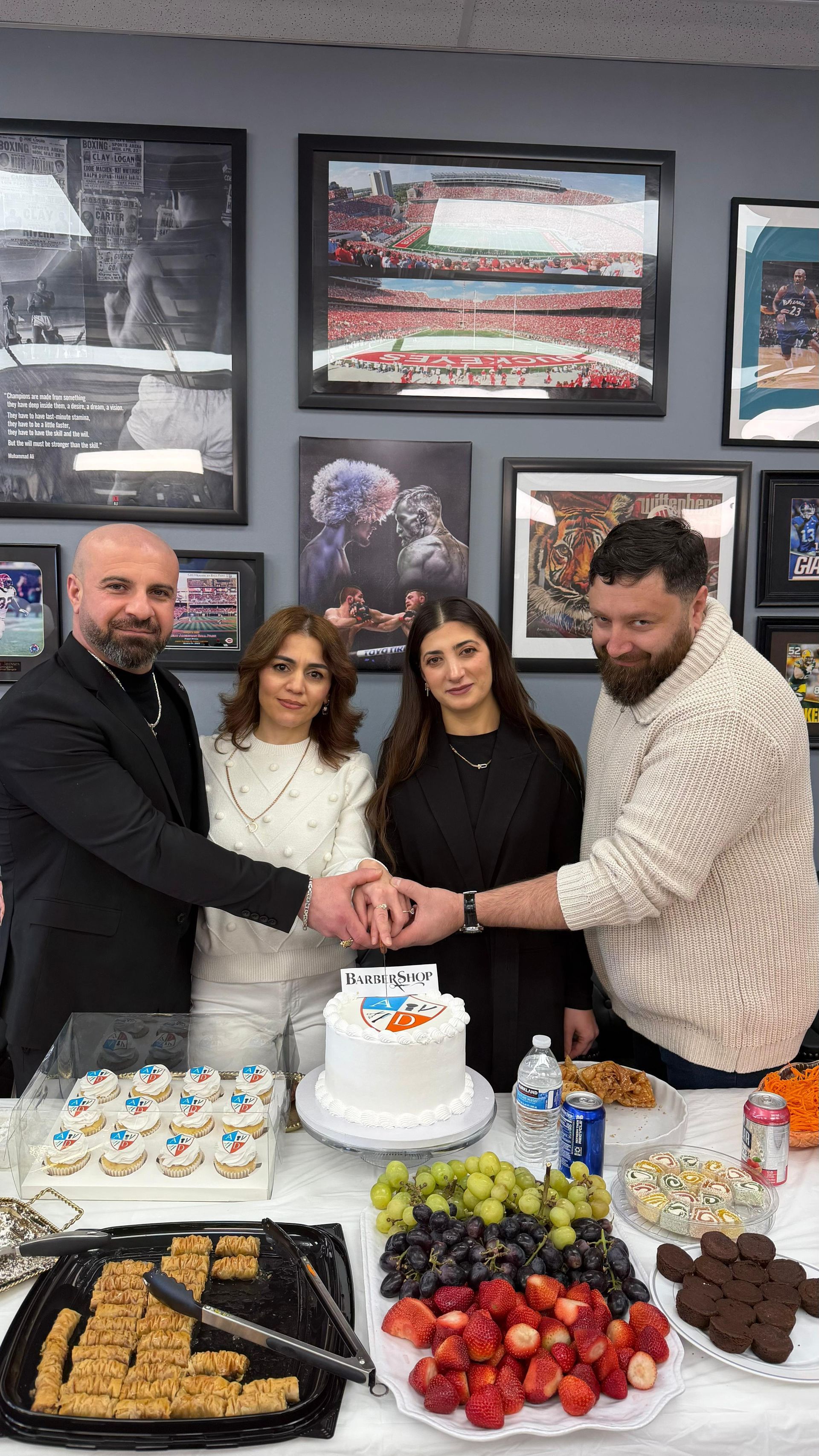 Four people cutting a white-frosted cake at a dessert table with pastries and fruit in an office setting