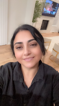 Smiling person taking a selfie indoors, with a TV and hanging plant in the background.