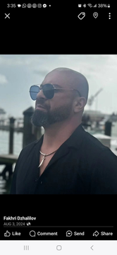 Man with sunglasses and beard in a dark shirt, standing outdoors by the water.