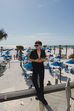 Person in black standing by a beach resort with blue umbrellas and palm trees under a clear sky