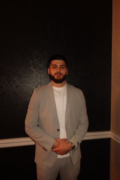 Man in a light gray blazer stands indoors against a dark wall, hands clasped, with a slight smile.
