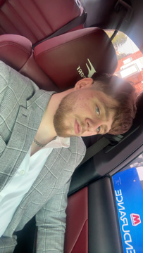 Man in a gray plaid blazer sitting in a car, taking a selfie with a slight smile.