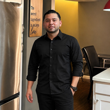Man in black shirt and pants standing in a modern indoor hallway near a kitchen and dining area