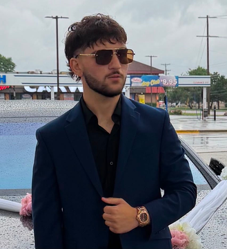 Man in navy suit and sunglasses standing outdoors beside a decorated car on a rainy day