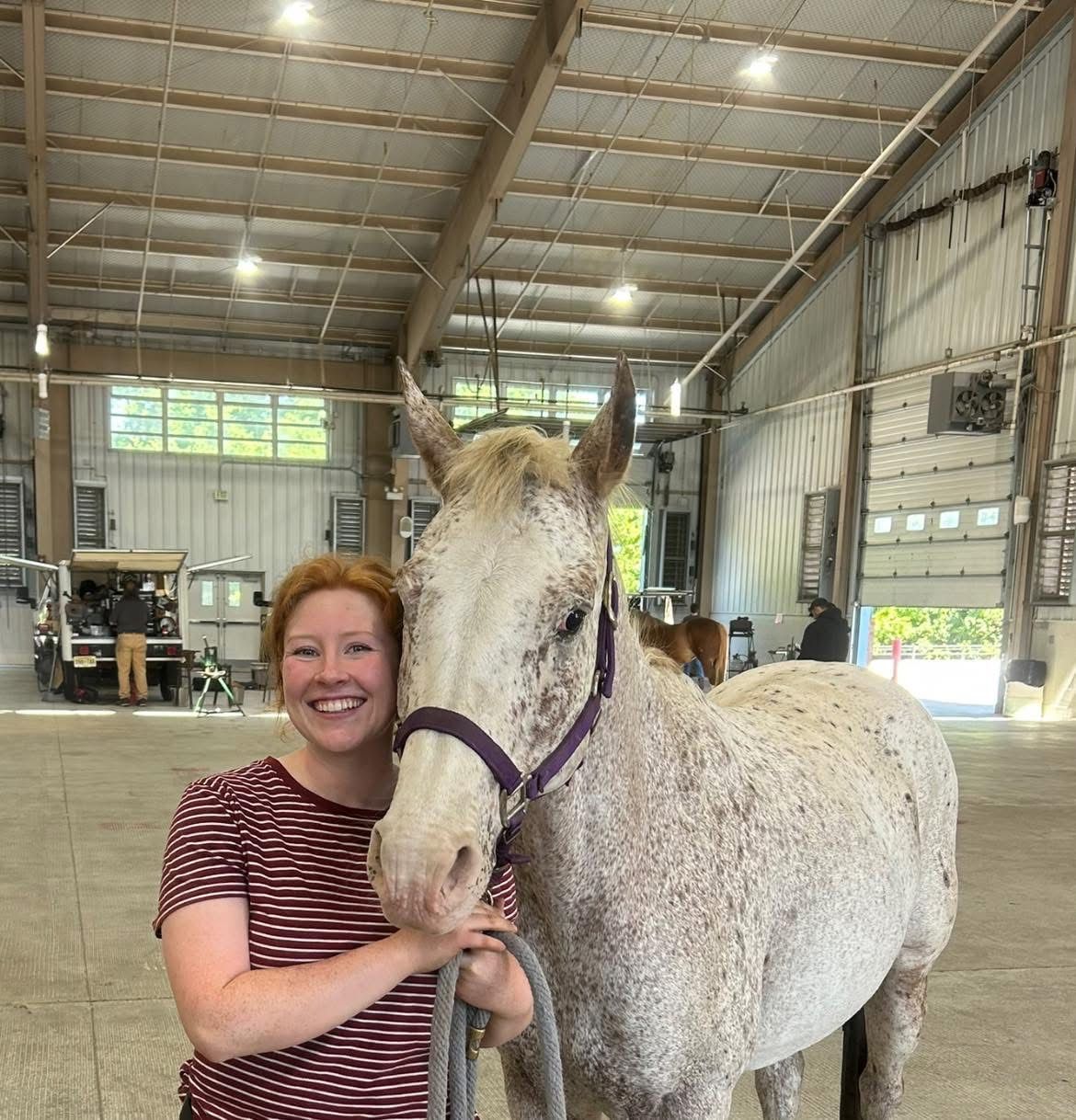 Smiling woman in striped shirt hugging a speckled horse in a barn.