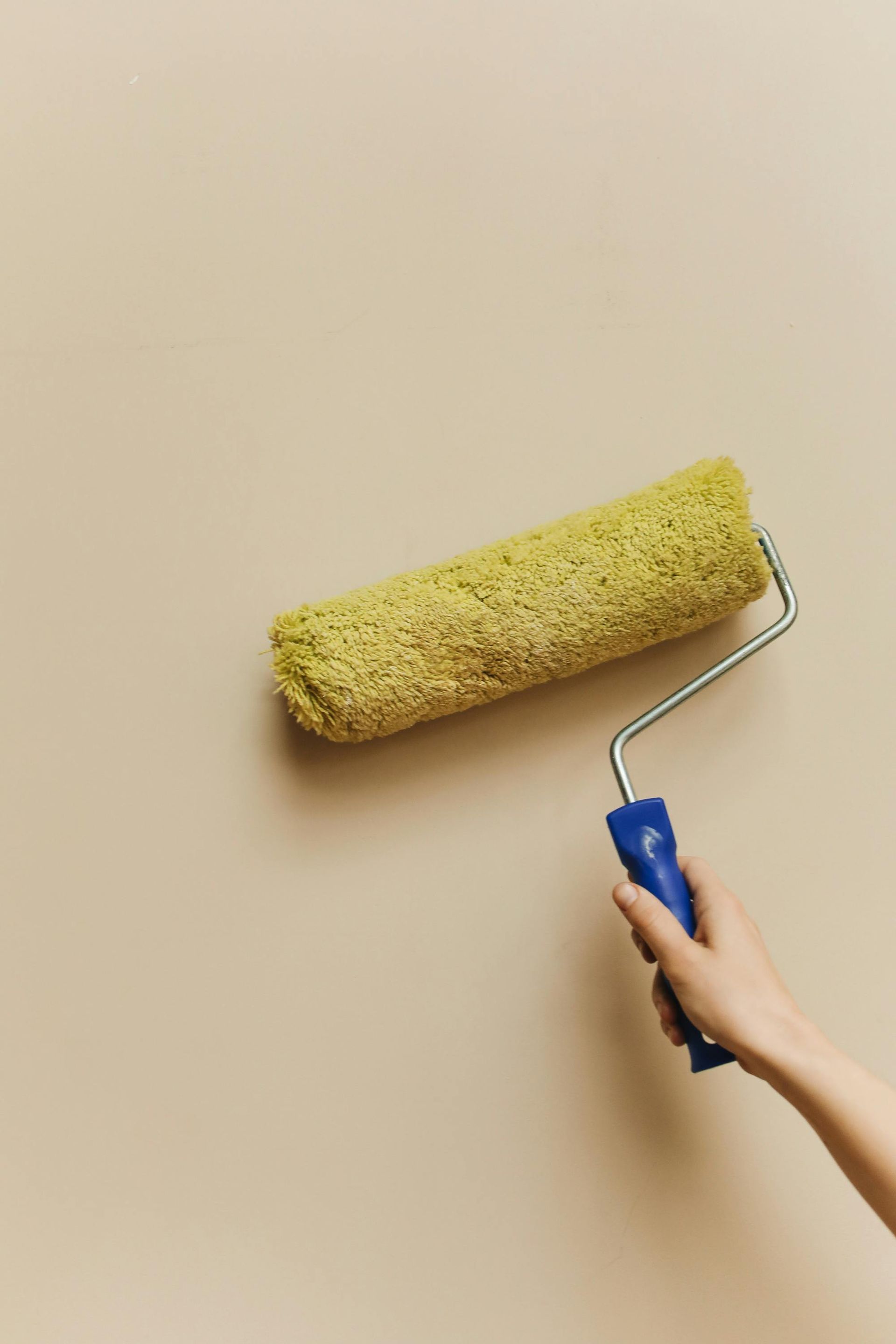Hand holding a paint roller applying yellow paint to a beige wall.