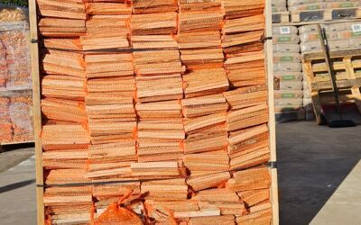 Una grande cassa di legno piena di sacchi di legna da ardere impilati e coperti da reti arancioni, all'aperto su un piazzale asfaltato.