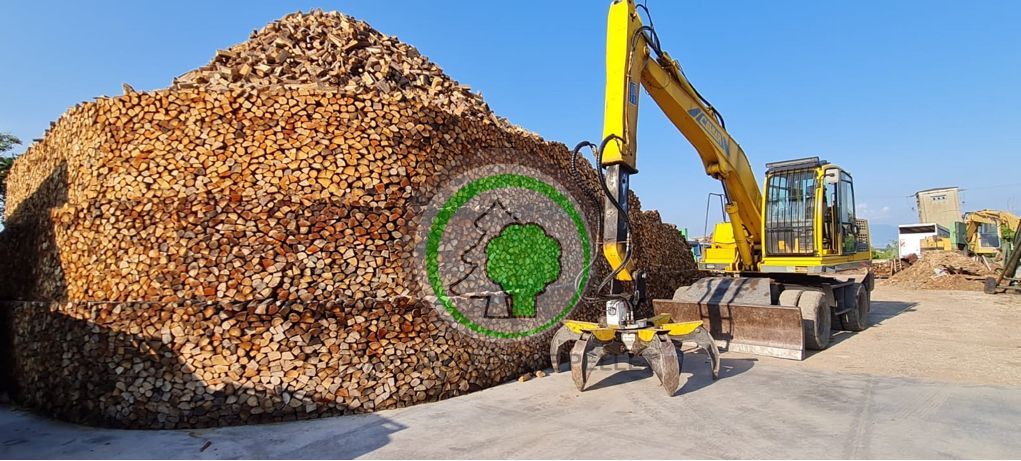 Un escavatore giallo si erge accanto a un'enorme catasta di trucioli di legno, su cui campeggia un logo circolare verde a forma di albero.