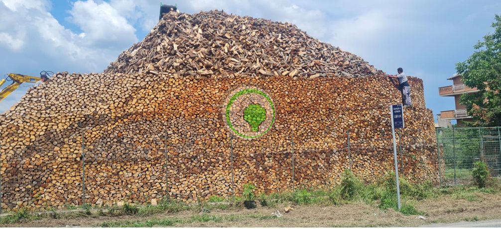 Un'enorme catasta di legna da ardere, simile a una montagna, accatastata in un campo, con un logo circolare verde ben visibile sulla parte anteriore.