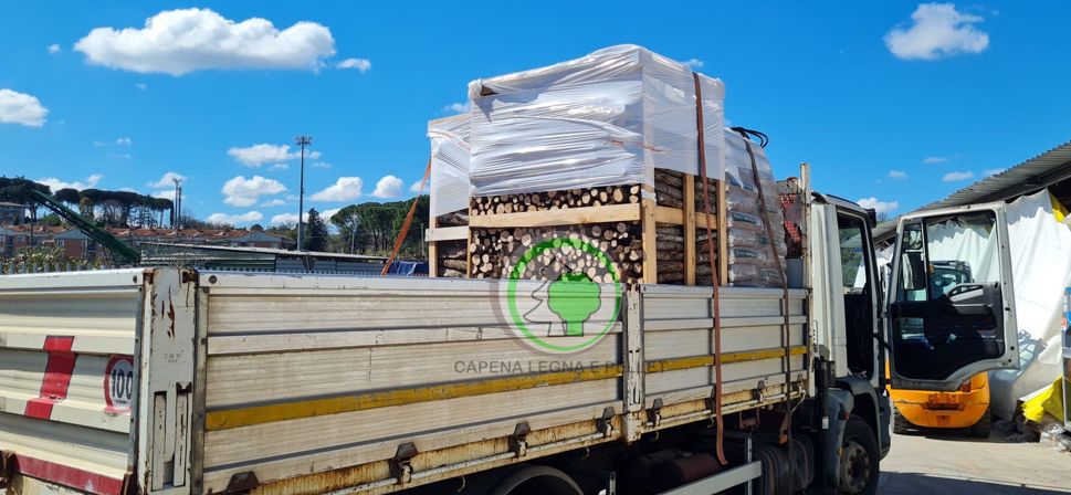 Un camion a pianale ribassato parcheggiato all'aperto, carico di grandi pallet industriali avvolti in pellicola bianca e fissati con cinghie arancioni.