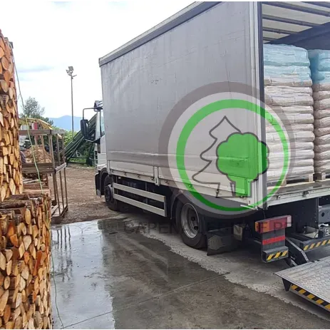 Un camioncino bianco per le consegne, carico di pellet di legno in sacchi, parcheggiato accanto a una grande catasta di legna da ardere in un cortile esterno.