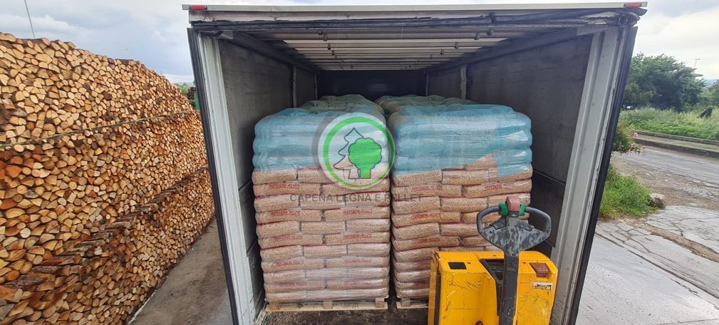 Un transpallet giallo è posizionato sul retro aperto di un rimorchio di un camion, carico di pile di merci insacchettate su pallet.