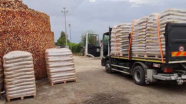 Un camion carico di merci insacchettate è parcheggiato accanto a una grande catasta di legna da ardere in un deposito di ghiaia.