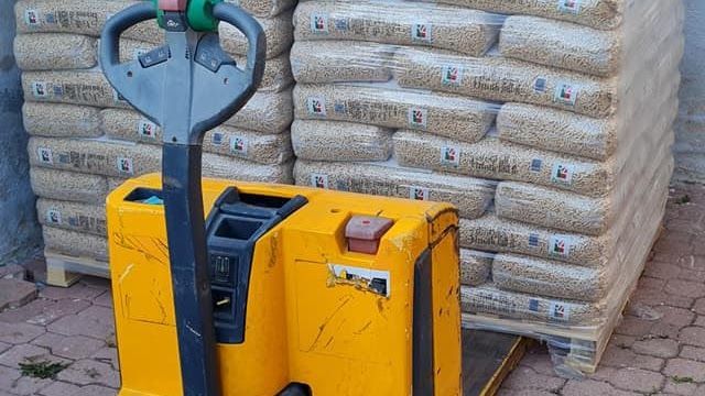 Un transpallet elettrico giallo si trova davanti a una pila di pallet con merci insacchettate di colore chiaro su una superficie esterna pavimentata.