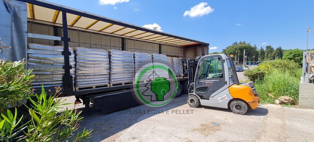 Un carrello elevatore parcheggiato accanto a un camion a pianale carico di pallet ordinatamente impilati di sacchi di pellet di legno, sotto un cielo azzurro brillante.