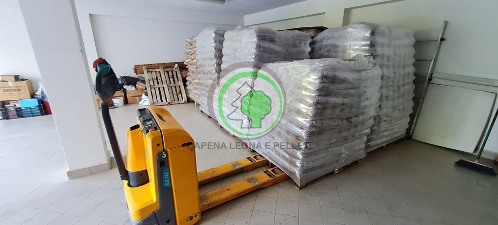 Un transpallet elettrico giallo si trova in un magazzino vicino a pile di sacchi avvolti su pallet di legno.