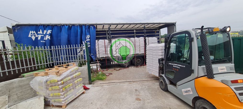 Un carrello elevatore trasporta un pallet di sacchi bianchi vicino al rimorchio di un semirimorchio in fase di carico in un'area industriale all'aperto.