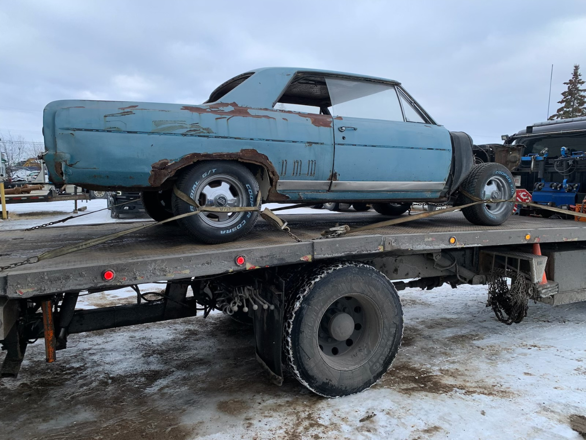 Rusty blue vintage car strapped on a flatbed trailer in a snowy outdoor lot