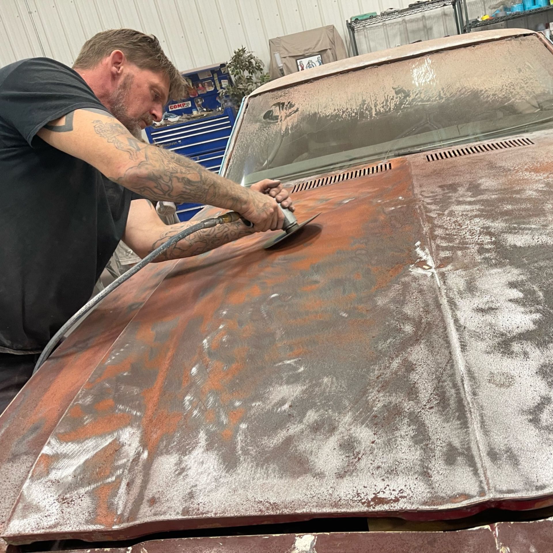 A man is sanding the hood of a rusty car.