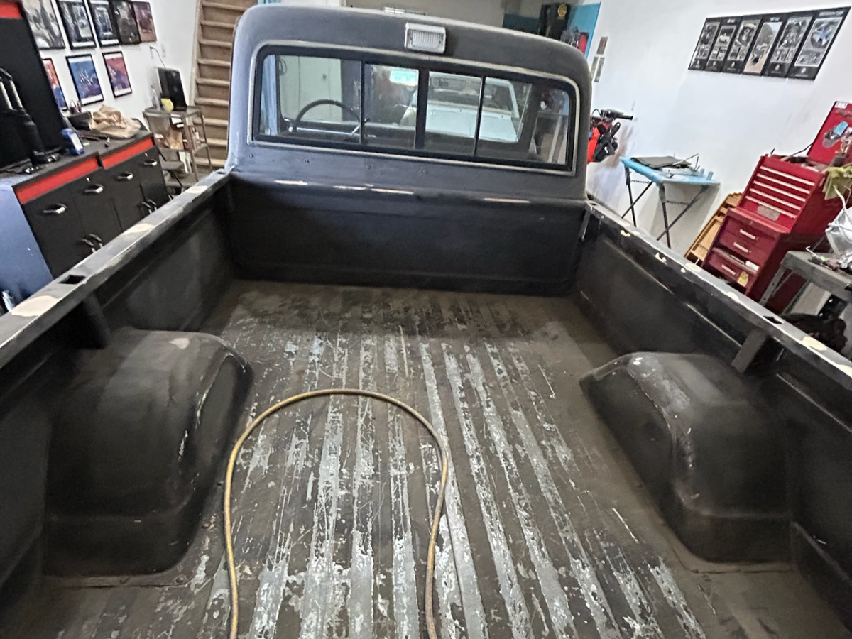 Interior of an old pickup truck bed in a workshop, showing worn metal floor and side panels.