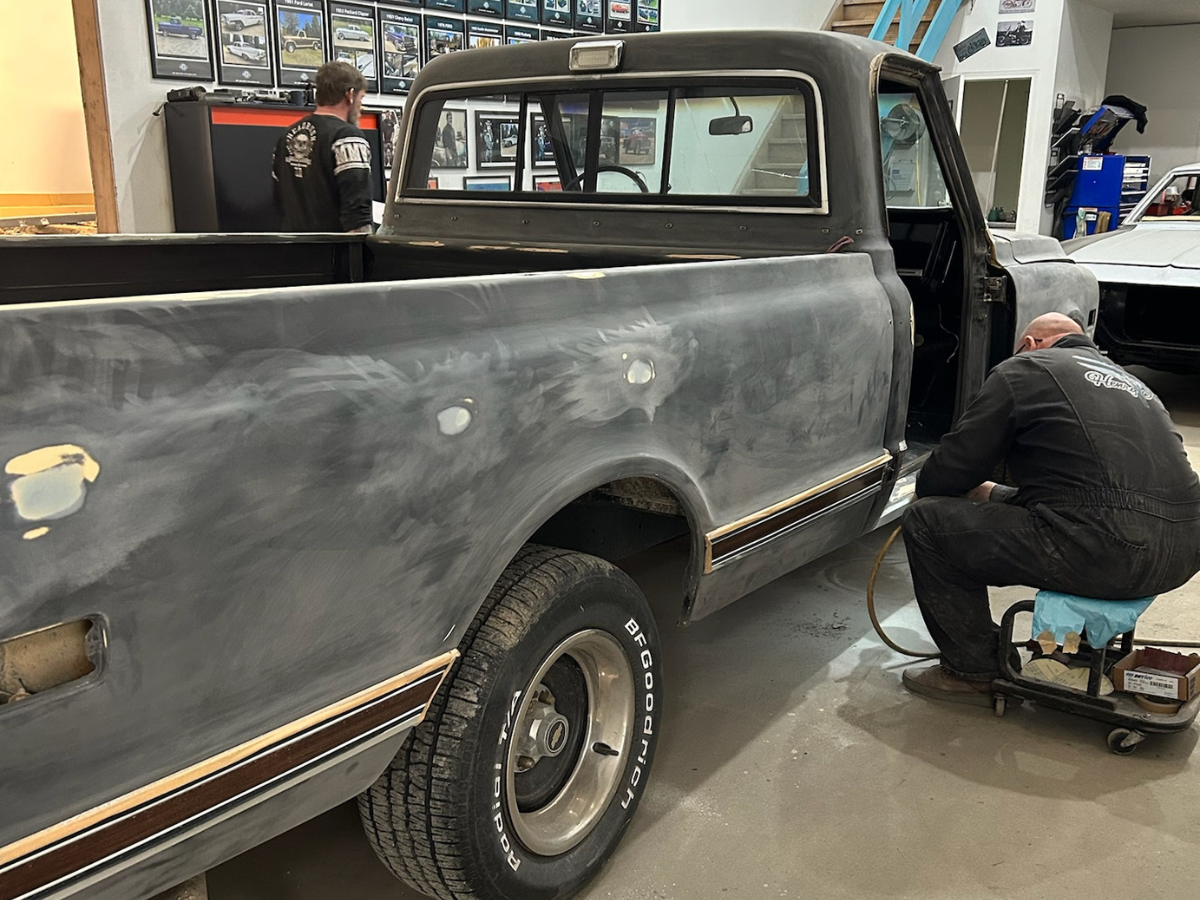 Matte black pickup truck in a garage, with a worker sanding the rear quarter panel on a stool